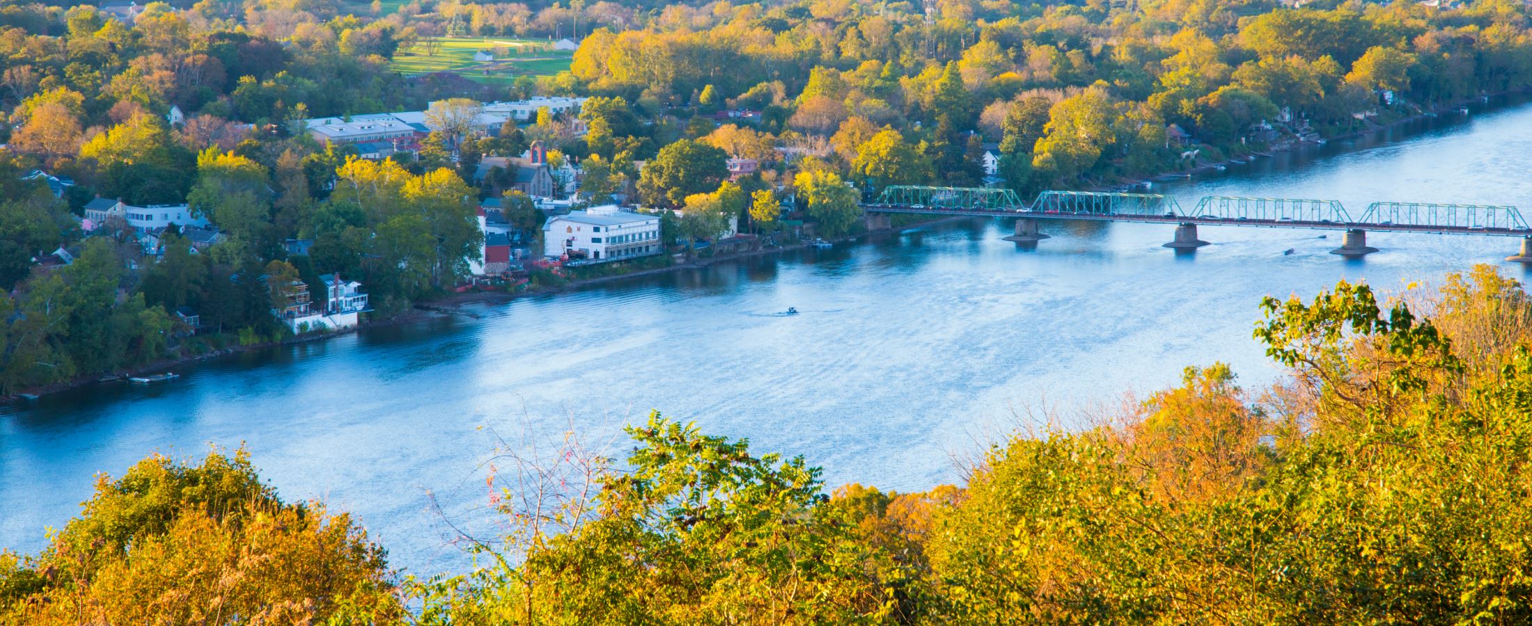 Overlook of a river crossed by a green truss bridge, with houses along the bank and autumn trees in the foreground